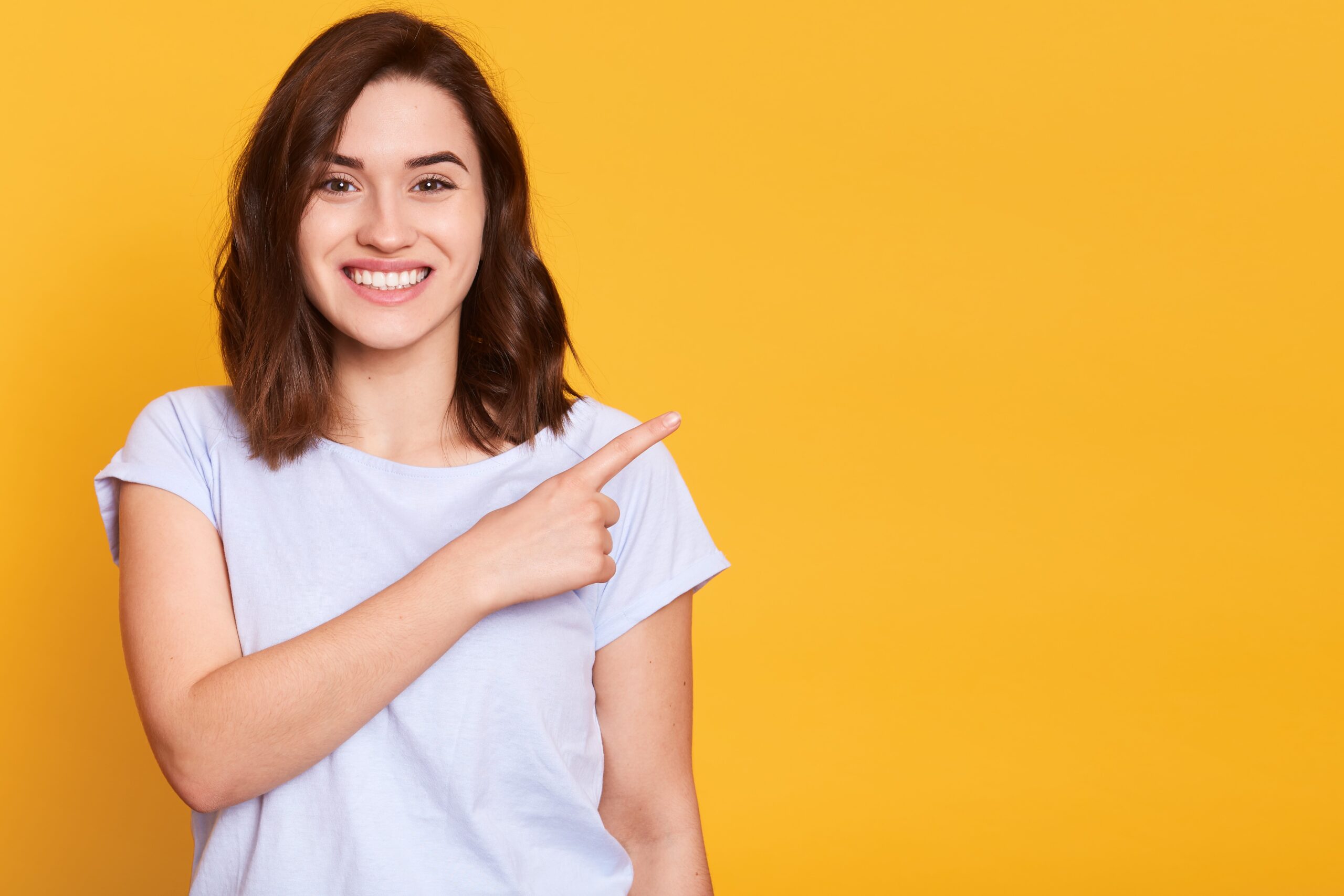 Close up portrait of nice cute adorable smiling charming cheerful girl pointing with her index finger aside, copy empty space for promotion text or advertisment, isolated over yellow background.